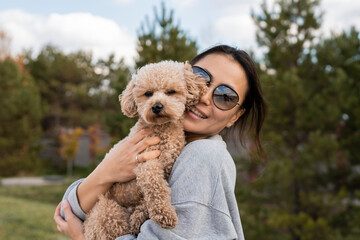 A young Asian woman holds her pet, a toy poodle, in her arms. Autumn walk.