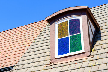 Close up of a dormer window with vibrant stained glass panels, set on a sloped roof covered with multicolored tiles, under a bright blue sky