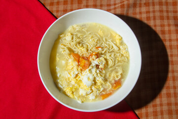 top view of mixed yellow noodle soup with egg and tomato in white bowl on red cloth on table