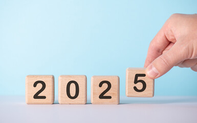 A hand adjusts wooden blocks with numbers to form the year 2025 against a light blue background,...