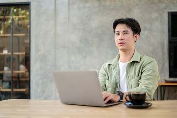 Fototapeta premium A handsome Asian man is sitting at a table in a modern loft coffee shop with his laptop.