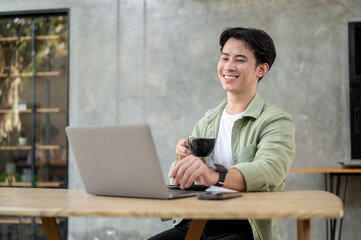 A handsome Asian man is working remotely from a coffee shop, enjoying coffee while working on laptop