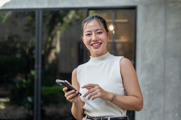 Beautiful Asian woman holding a smartphone in her hand, standing outdoors and smiling at the camera.