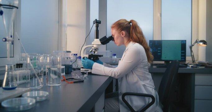 Young female scientist sits in chair, looks under microscope and examines samples of experimental drugs in medical science laboratory. Innovations in pharmaceutics and pharmacology. Medicine concept.