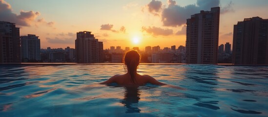 A woman swims in a rooftop infinity pool while watching the sunset over a city skyline.
