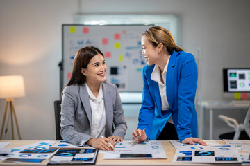 Two women in suits are talking at a desk with a white board behind them