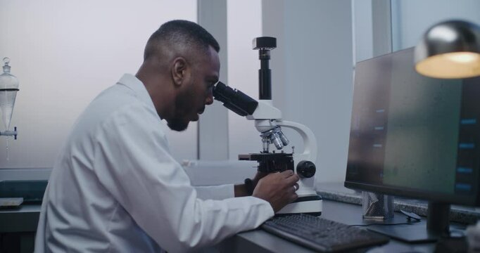 Medical science laboratory: African American microbiologist looks under microscope, does analysis of test sample using high-tech equipment. PC screen shows image of cells or bacteria. Microbiology.