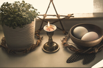 Home pagan altar on windowsill with bowl with eggs, pentacle, the first greens in the pot, candle and birds feathers under the sun's rays, selected focus.