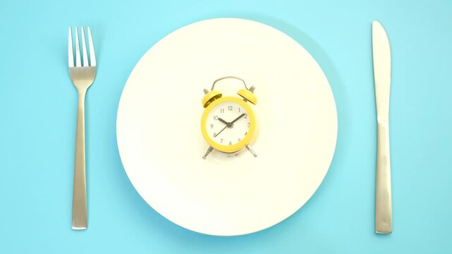 Empty plate with alarm clock on blue background, intermittent fasting concept.