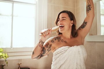 Woman, happy and brush in bathroom for singing, morning routine and excited for self care with towel. Female person, smile and dance in home for personal hygiene, grooming and weekend with tattoo