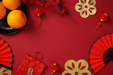 Chinese New Year flat lay composition with traditional decorations: oranges on a black plate, red paper fans, gold cutouts, flowers, and red lanterns on red background. Lunar New Year 2025 card.