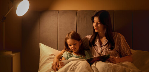 Happy mother and her little daughter reading fairy-tale in bed at night