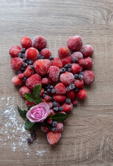 Top view of frozen strawberries and blueberries on a wooden table, arranged with green leaves and a pink rose in the shape of a heart. Elegant and vibrant presentation.