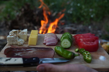 A board with delicacies arranged on it for eating. A fire is burning in the background. Tomato, cucumber, meat, cheese, yellow cheese, hot pepper, cutting knife.