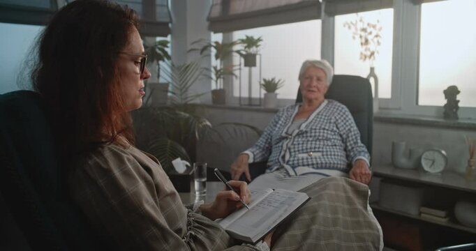 Female psychologist listens to elderly client during appointment, asks clarifying questions, writes down woman's complaints about anxiety and depression in notebook. Psychology. Work of psychologist.