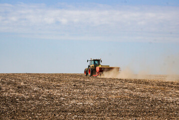 Fototapeta premium The tractor moves along the field and applies the fertilizer to the soil. Clouds of dust rise from the tractor trailer with dry soil in its wake.