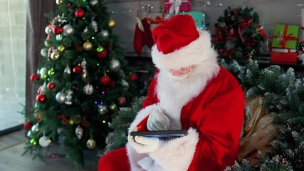 Busy Santa with tablet and stylus making list of gifts and Christmas chores sitting near Christmas tree and festive garlands. 