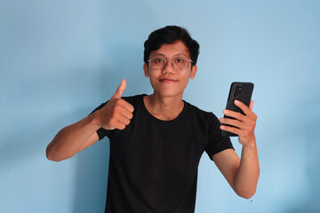 young Asian man in black t-shirt holding mobile phone and making thumb up gesture, approve good thing