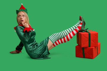 Emotional young woman in elf costume with Christmas gift boxes on green background