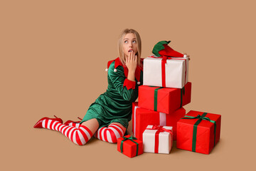 Shocked young woman in elf costume with Christmas gift boxes on brown background