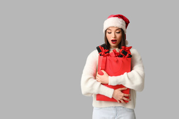 Young woman in Santa hat holding shopping bag with gift boxes on grey background