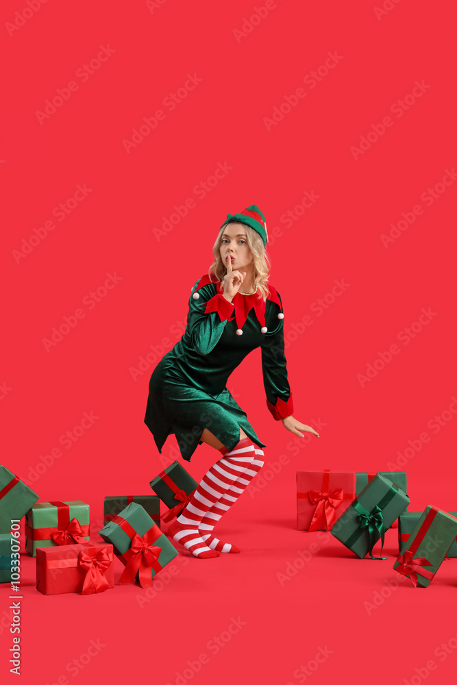 Wall mural Beautiful young woman dressed as elf with Christmas gifts showing silence gesture on red background