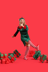Happy young woman dressed as elf with Christmas gifts on red background