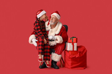 Cute little girl with gift boxes telling Santa Claus her wish on armchair against red background