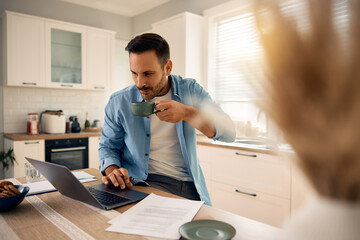 Smiling man drinking coffee while working on laptop at home.