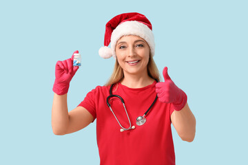 Mature female doctor in Santa hat with jar of pills showing thumb-up gesture on blue background