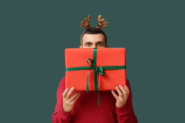 Young man in reindeer horns with Christmas gift box on green background