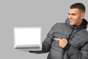 Young man in stylish puffer jacket with modern laptop on grey background