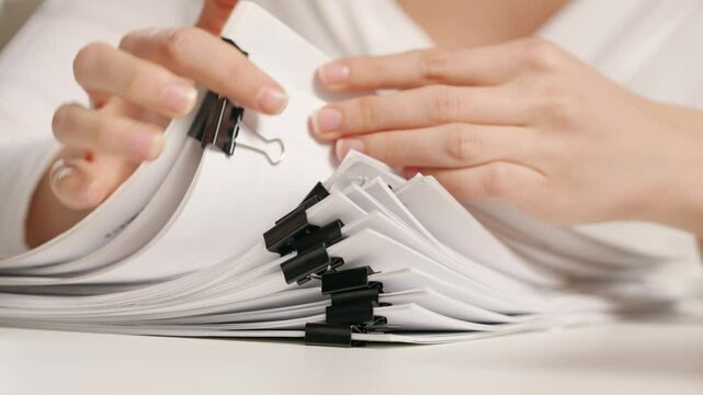 Woman sitting at office desk searching through overload stack of paper documents with binder clips., close up Bureaucracy business concept.