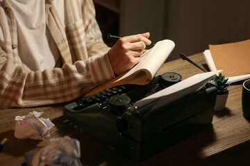 Young writer with notebook and vintage typewriter working at table in room, closeup