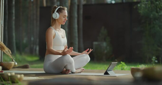 Seated Peacefully in a Serene Nature Setting, Beautiful Woman Wears Headphones while Meditating with the Aid of a Tablet Computer App. She Focuses on Guided Meditation, Finding Inner Balance and Calm