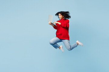 Obraz premium Full body little kid teen IT girl of African American ethnicity wear red sweatshirt jump high hold use work on laptop pc computer isolated on pastel plain blue background. Childhood lifestyle concept.