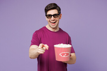 Young Caucasian man wear violet t-shirt casual clothes 3d glasses watch movie film hold bucket of popcorn in cinema point index finger camera on you isolated on pastel light purple background studio.