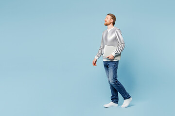 Full body side view young happy IT man he wear striped sweatshirt casual clothes hold closed laptop pc computer walk go isolated on plain pastel light blue cyan background studio. Lifestyle concept.