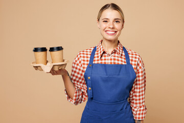 Young happy woman barista bartender barman wear blue apron red shirt work in coffee shop hold takeaway delivery paper cup tea to go isolated on plain beige background. Small business startup concept.