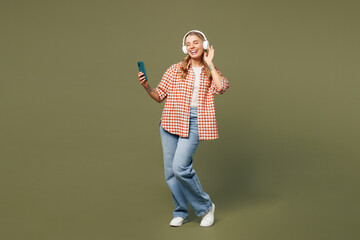 Full body young woman wear red shirt white t-shirt casual clothes listen to music in headphones hold in hand use mobile cell phone isolated on plain pastel green background studio. Lifestyle concept.