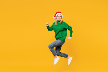 Obraz premium Full body merry fun young woman wears green casual sweater Santa hat posing jump high run fast look camera isolated on plain yellow background. Happy New Year, celebration Christmas holiday concept.