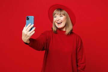 Elderly blonde woman 50s years old she wear sweater hat casual clothes doing selfie shot on mobile cell phone post photo on social network isolated on plain red background studio. Lifestyle concept.