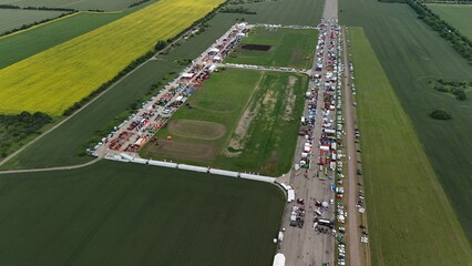 This aerial image shows a large agricultural event or trade fair, amidst expansive fields. Multiple rows of tents, machinery, and equipment are neatly arranged on a long track, attracting numerous