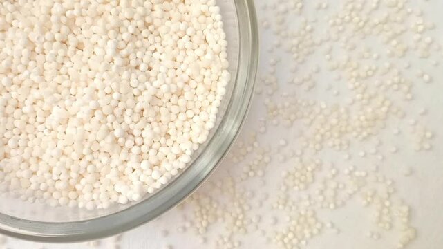 Sago pearls or tapioca pearls in a glass bowl rotating top left corner with spices spilled around it, white background, closeup, herbs and spices