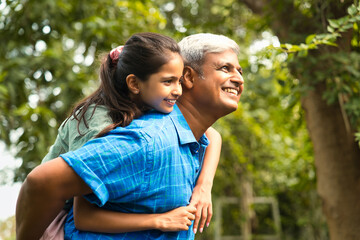 Fototapeta premium Indian father in his forties joyfully carrying daughter on his back for a piggyback ride outdoors