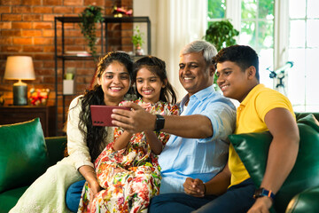 Indian family of four smiling together, wife capturing a selfie on her smartphone indoors