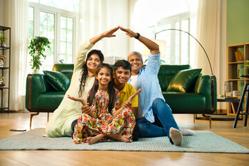 Indian asian young family of four sitting on carpet, parents creating roof sign at the camera