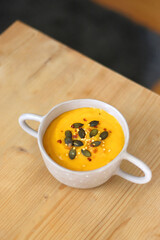 Bowl of creamy pumpkin and potato soup, with pumpkin seeds and chilli flakes toppings. Selective focus, wooden background.