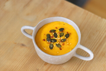 Bowl of creamy pumpkin and potato soup, with pumpkin seeds and chilli flakes toppings. Selective focus, wooden background.
