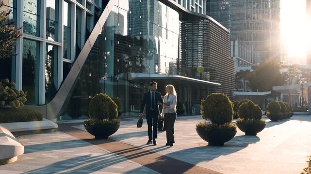 Two Business People Talking On the Go Outside Office Building Entrance with Sun Setting In Backdrop - wide slow motion. Businesspeople having a discussion while walking in the city